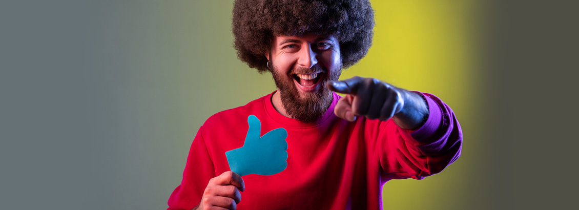 Hombre Joven Sonriendo Mientras Sostiene Ícono de Me Gusta y Señala la Cámara con su Mano